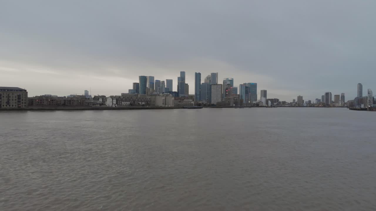 Plano de drone com dolly para frente e ascensão sobre Canary Wharf, Ilha dos Cães, Londres, sobre o rio Tâmisa.