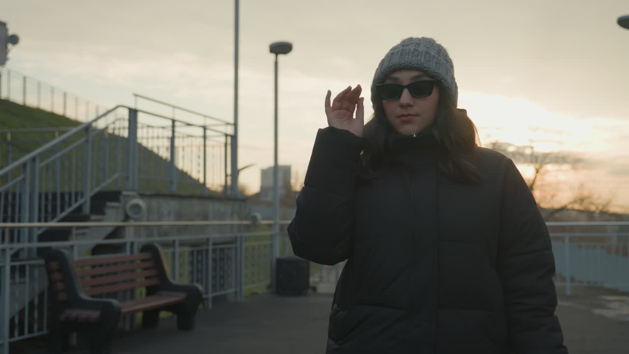 Rooftop walkway woman walking bag, wool hat, sunglasses, brisk confident steps, horizon glow, bench and railing framing, subtle hand gesture as if modeling, cinematic warm tones and shallow focus