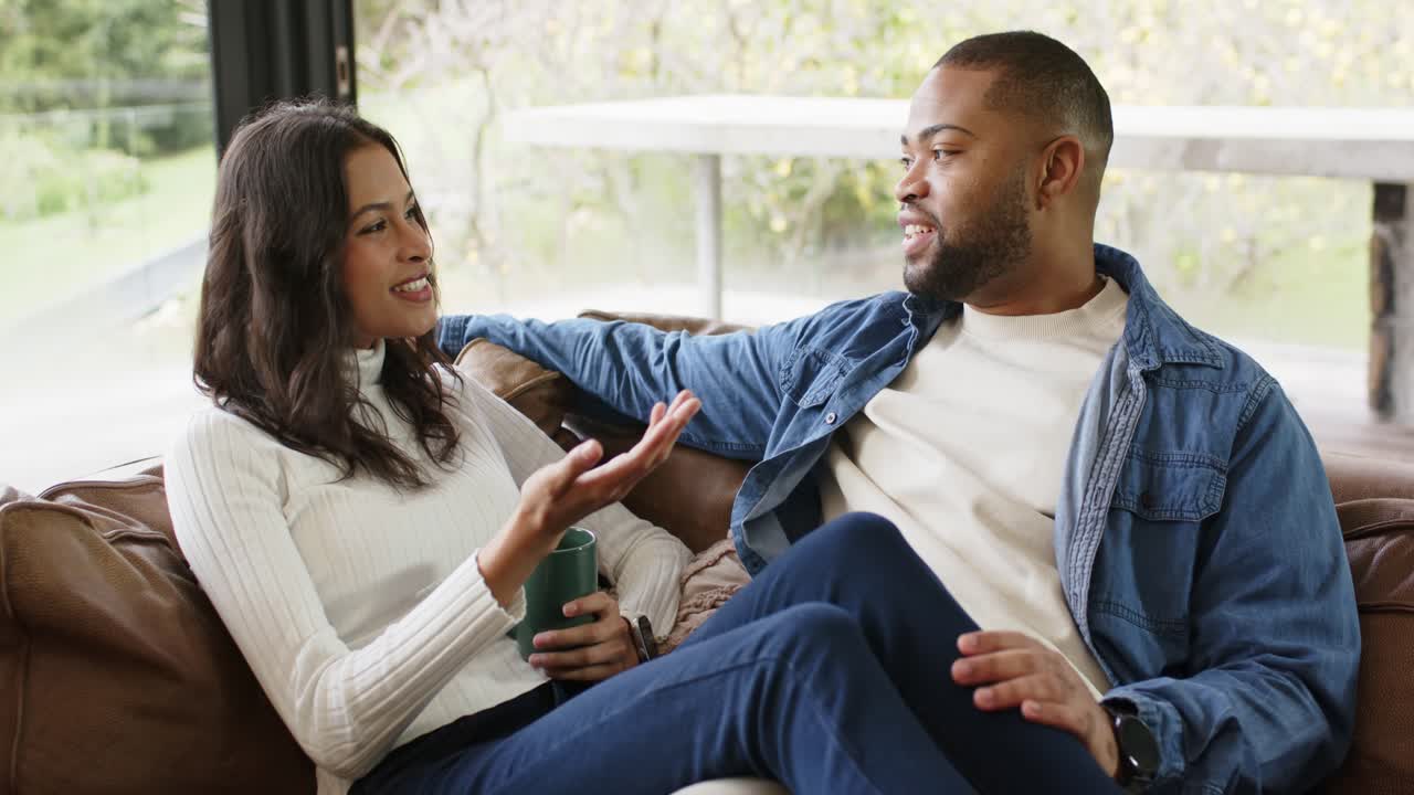 After sipping green mug, diverse couple exchanging ideas and gesturing on leather sofa by window