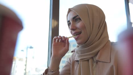 beautiful muslim woman in headscarf and fashionable modern trendy clothes eats potatoes fries in restaurant with happy smiling