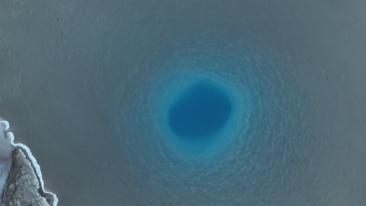 아이슬란드의 상징적인 게이시르 온천을 드론으로 촬영하여 활기찬 파란색 중심과 주변 지열 지형을 보여줍니다.