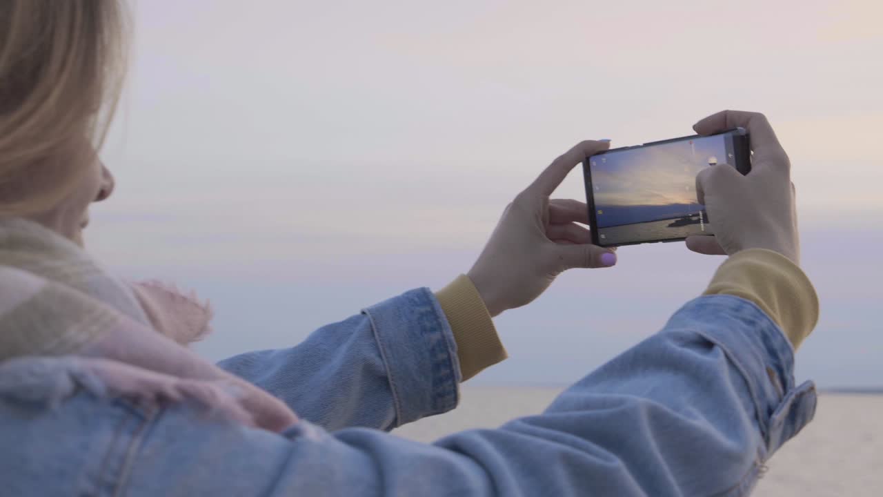 las manos femeninas con el teléfono hacen la foto del atardecer en la orilla del mar.