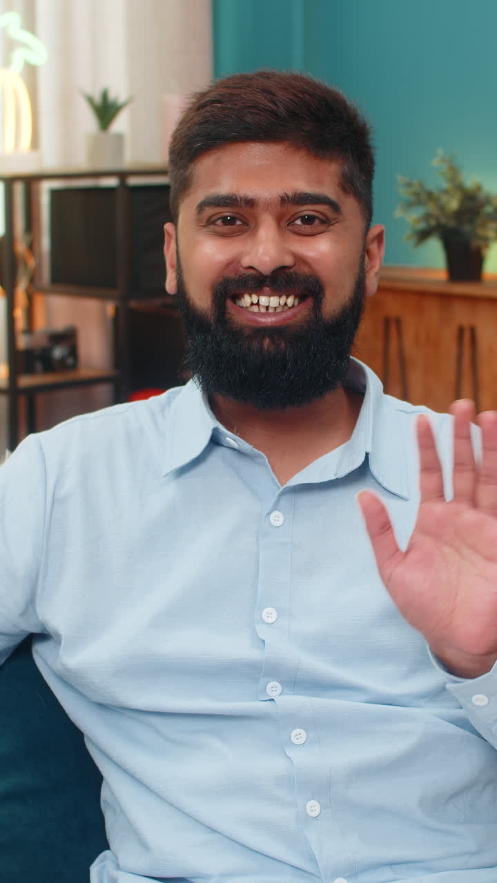 Indian man waves camera smile friendly hello gesture shows connection smartphone video chat indoors