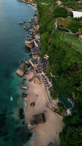 a vertical drone view captures rock bar built on large coastal rocks as people enjoy dinner near the blue ocean surrounded by natural stone and relaxing seaside ambiance in bali.