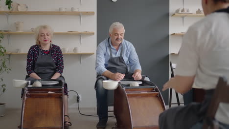 medium shot of middle aged ceramic artist teaching group elderly caucasian woman and senior man how to wedge clay sitting at desk in art studio. people enjoying talking at work