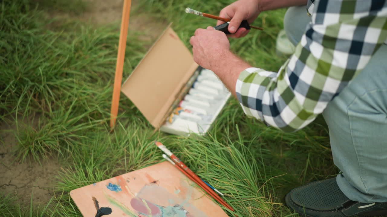 un dibujante en una camisa a cuadros se inclina en un campo de hierba, abriendo cuidadosamente un tubo de pintura de una caja de pinturas. una paleta con varios colores también está en la hierba