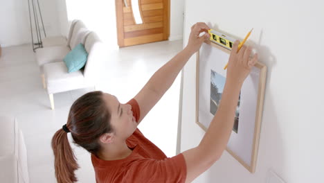 focused plus size biracial woman using spirit level hanging picture in living room, slow motion