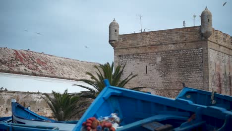 Camera-slides-right-with-the-fortress-of-Essaouira,-Morocco-standing-tall-over-the-fishing-boats-in-the-port-area