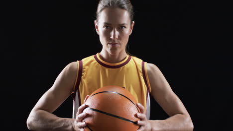 Una-Joven-Caucásica-Posa-Con-Confianza-En-Un-Uniforme-De-Baloncesto,-Sosteniendo-Una-Pelota-Sobre-Un-Fondo-Negro.