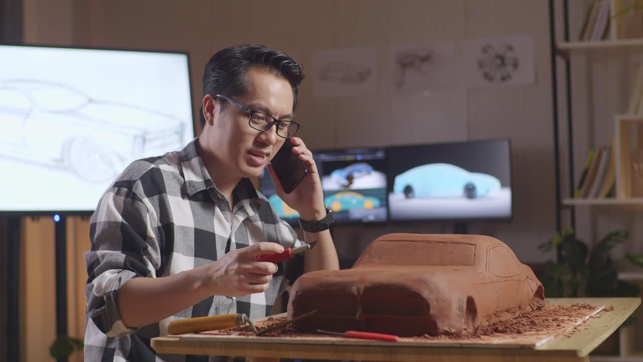 hombre asiático diseñador de automóviles hablando en su teléfono inteligente mientras usa rastrillo o alambre para suavizar la superficie y crear detalles en la escultura de arcilla de automóvil en el estudio