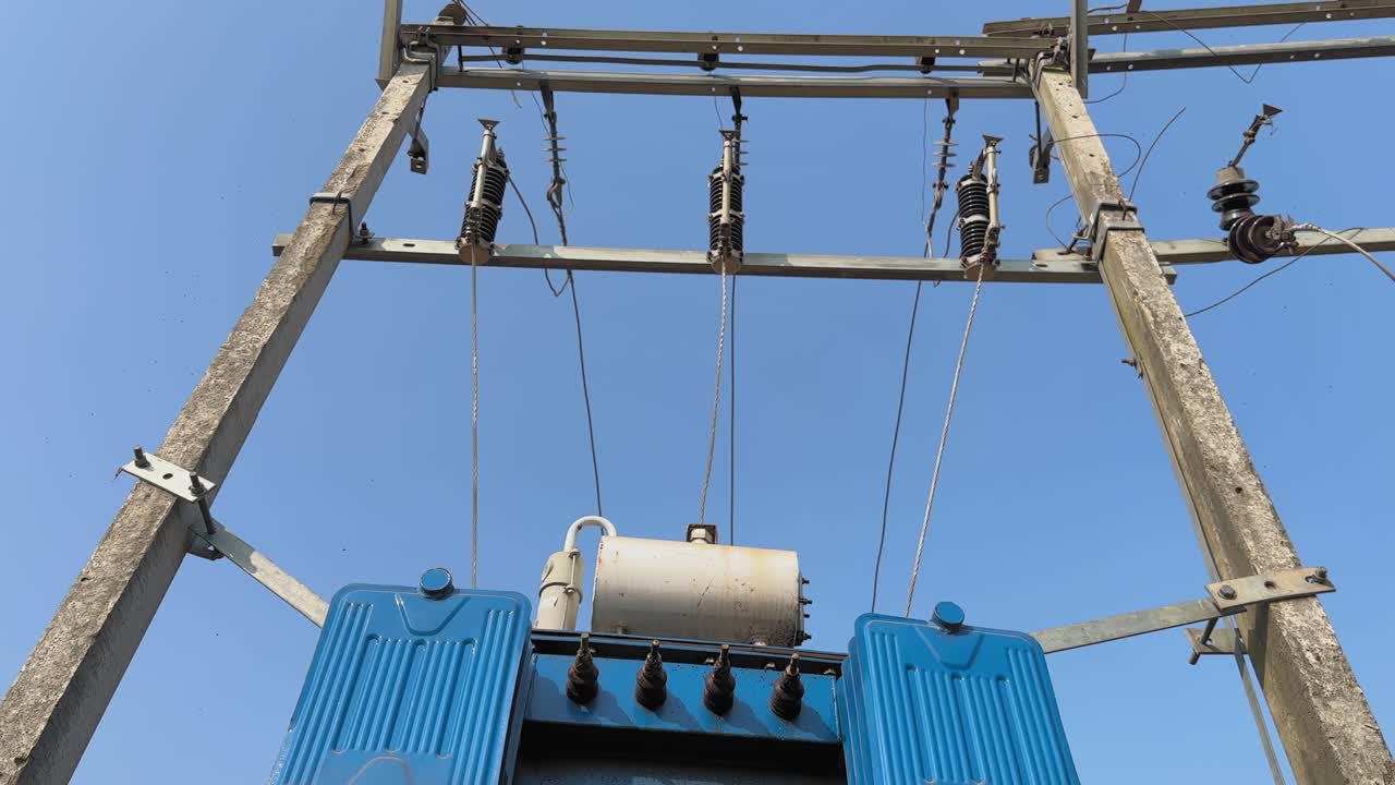 Inquadratura dal basso verso l'alto dalla base di un palo della luce fino al trasformatore elettrico e agli isolatori, linee elettriche che si estendono verso l'alto contro un cielo azzurro limpido, enfatizzando la scala e l'infrastruttura