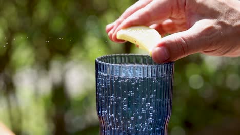 A hand squeezes lemon juice into a textured blue glass, capturing droplets in motion against a blurred green background.