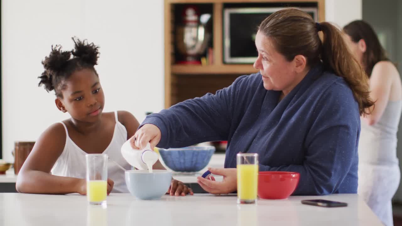 Happy caucasian lesbian couple and their african american daughter eating breakfast in kitchen