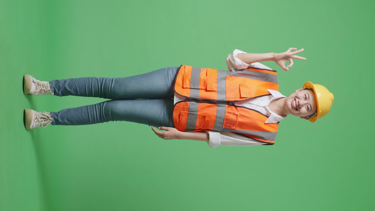 안전 헬을 입은 아시아 여성 엔지니어의 몸 전체가 미소 짓고 녹색 화면 배경 스튜디오에 서있는 동안 카메라에 은 몸짓을 보여줍니다.