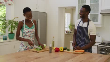 una pareja afroamericana sonriente con delantales hablando y preparando comida en la cocina