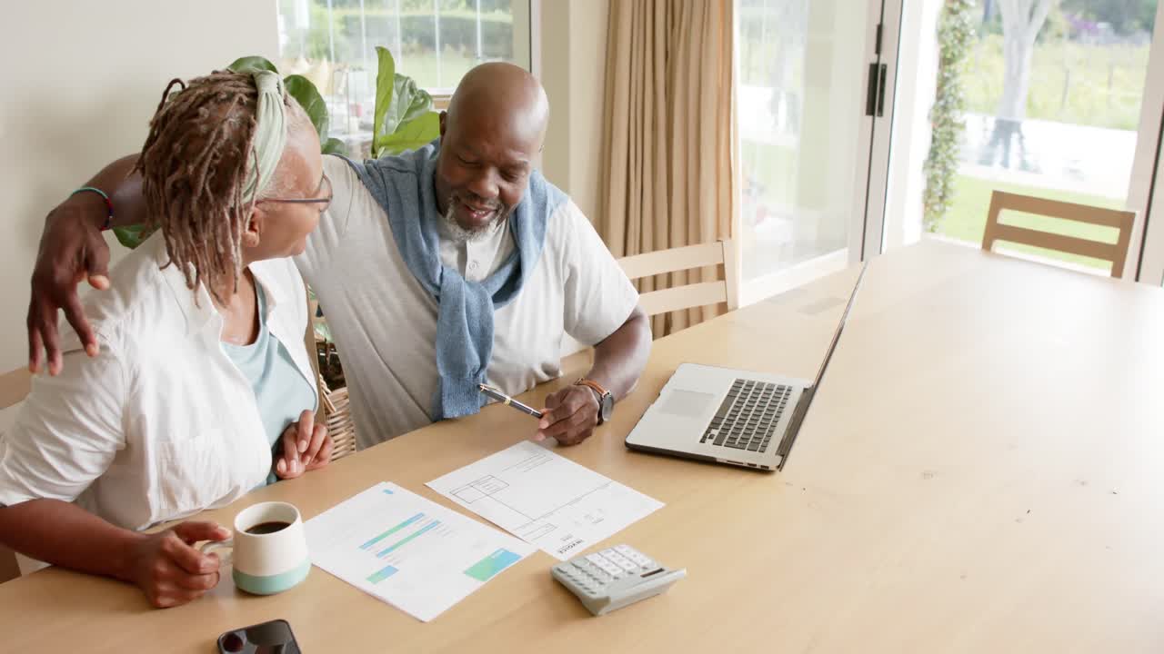 Happy african american senior couple doing paperwork using laptop at home, slow motion