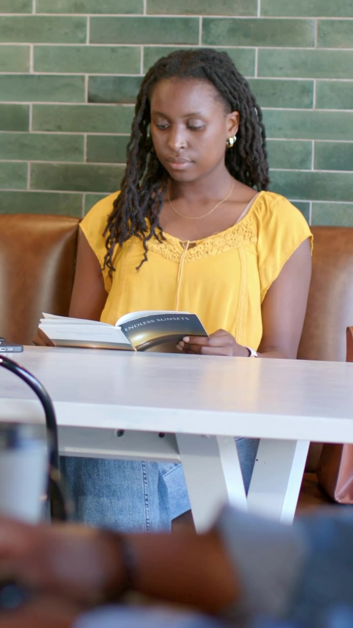 Vertical video: Studying woman with yellow blouse holding paperback and turning pages at cafe booth