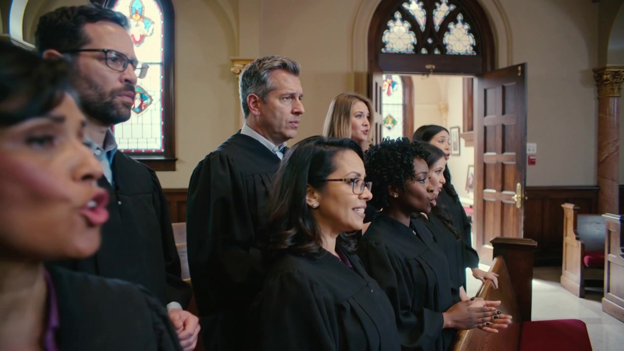 La cámara retrocede, revelando a los miembros del coro cantando en la iglesia durante el culto, vistiendo túnicas negras.