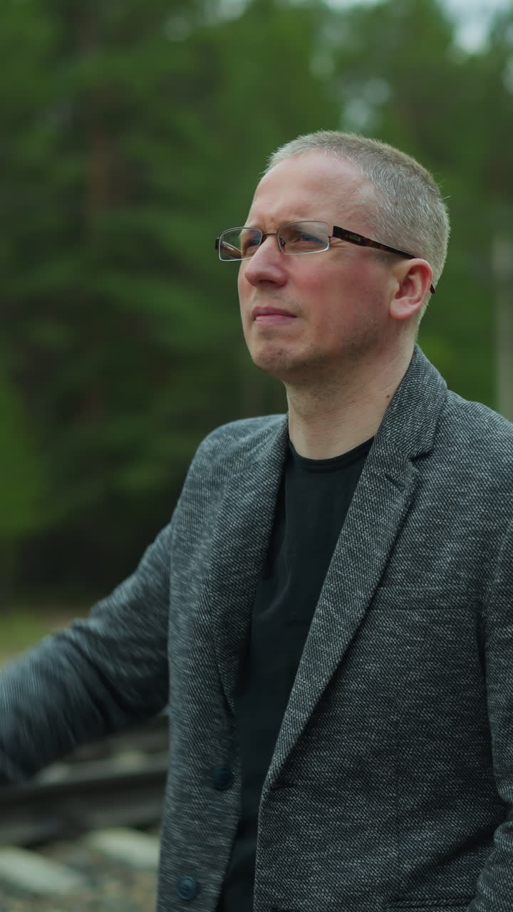 Close-up view of a man in glasses and a gray jacket standing on a railway track, looking worried as he rubs his head with one hand