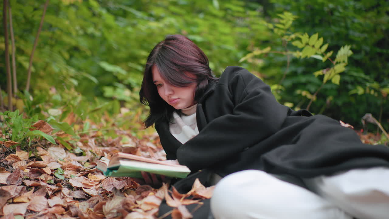 Forest visitor lying on leaf carpet, focused on open book, flipping pages attentively, cozy in black coat and striped sweater, calm mood, quiet autumn park scene with rich foliage