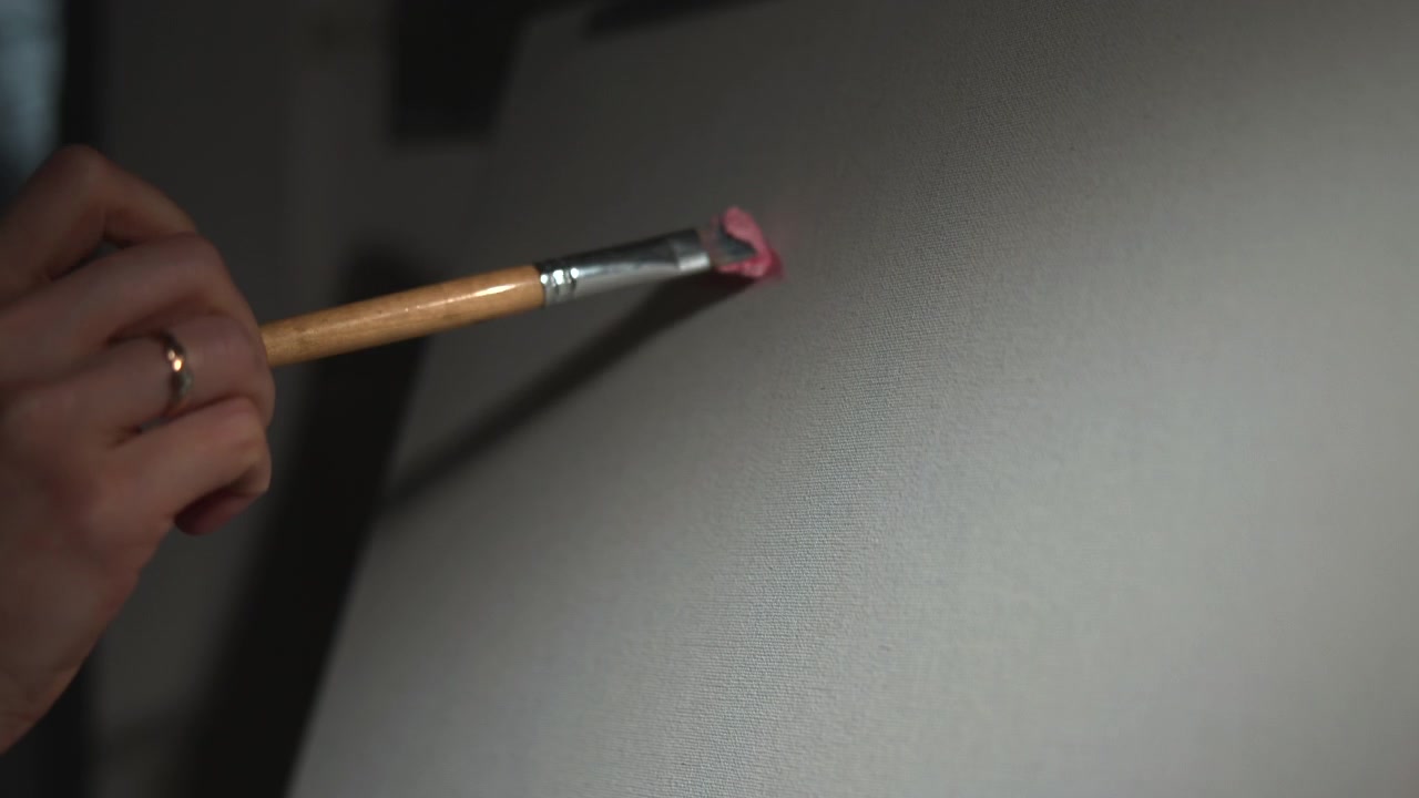Close-up of a small red brush on a white handle touching a plain light gray wall, with soft indoor lighting and a dark vertical object blurred in the background