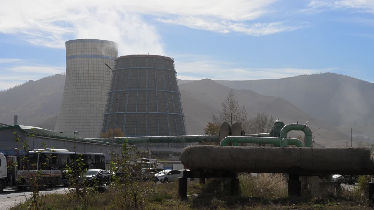 Grandi tubature di riscaldamento corrono accanto a una centrale termica a Ulaanbaatar, in Mongolia. Il fumo si alza denso dalle torri di raffreddamento dietro una recinzione metallica, evidenziando la portata dell'infrastruttura energetica urbana.