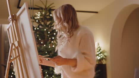 blonde girl painting on canvas in bedroom with christmas tree in the background