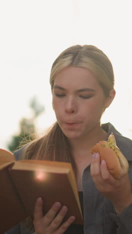 dama comiendo un bocadillo mientras lee un libro al aire libre, sentada en un banco del parque rodeada de vegetación, con árboles borrosos y rayos de luz solar que se reflejan suavemente en el fondo