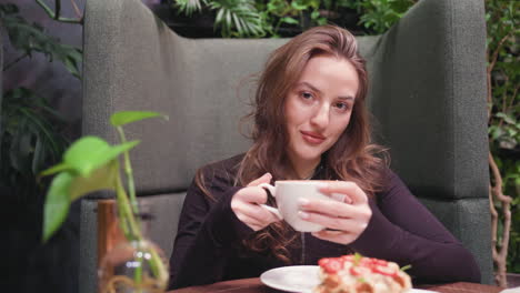close view of young woman sitting on cozy couch holding tea cup with calm expression, surrounded by indoor plants and soft lighting, dessert plate on table with decorative flower in glass vase