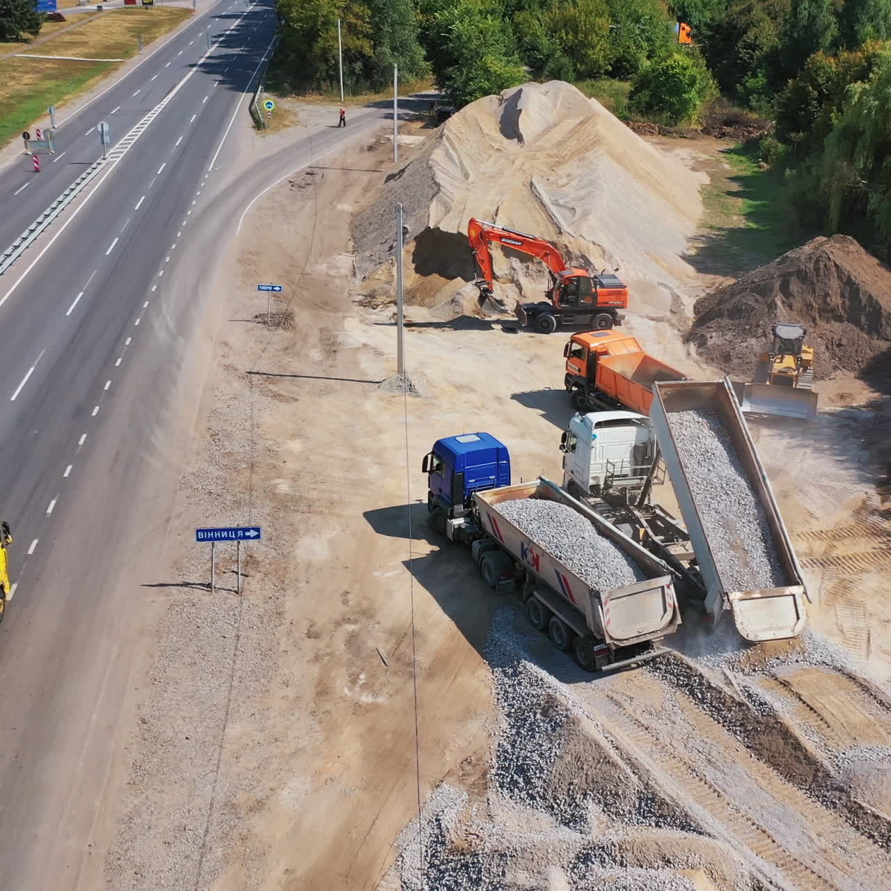 Trucks pouring out granite stone. Heavy machinery carry metal for construction new asphalt road near the highway. Aerial view.