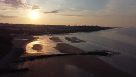 Sunset-rays-beam-fiery-golden-colours-across-relaxing-beach-ocean-coastline-aerial-view-slow-pull-back