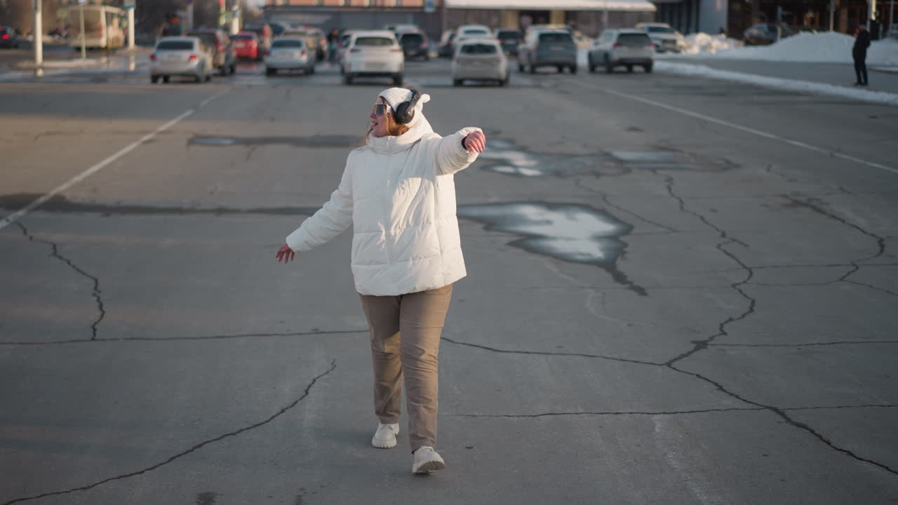 Young woman in winter jacket swaying body rhythmically on cracked pavement, surrounded by urban traffic and water patches, wearing headphones and sunglasses, expressing happiness in cold atmosphere