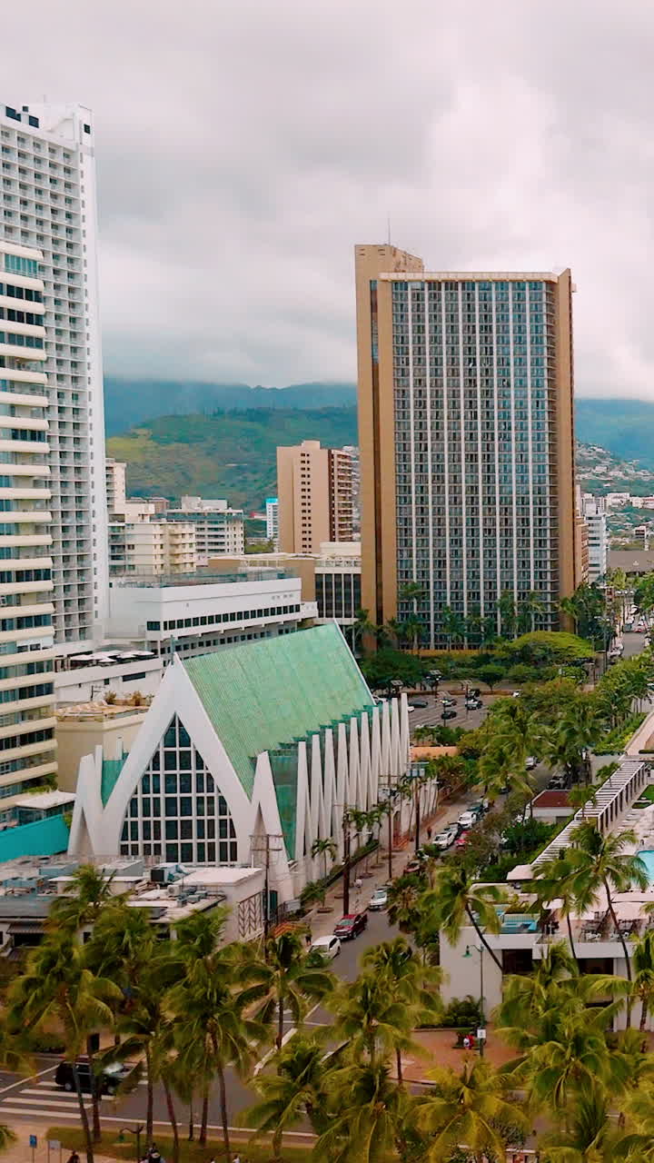 Vista elevata di una spiaggia sabbiosa con persone, palme ed edifici a molti piani. Scena di spiaggia urbana con architettura moderna e una cappella bianca. Video verticale.