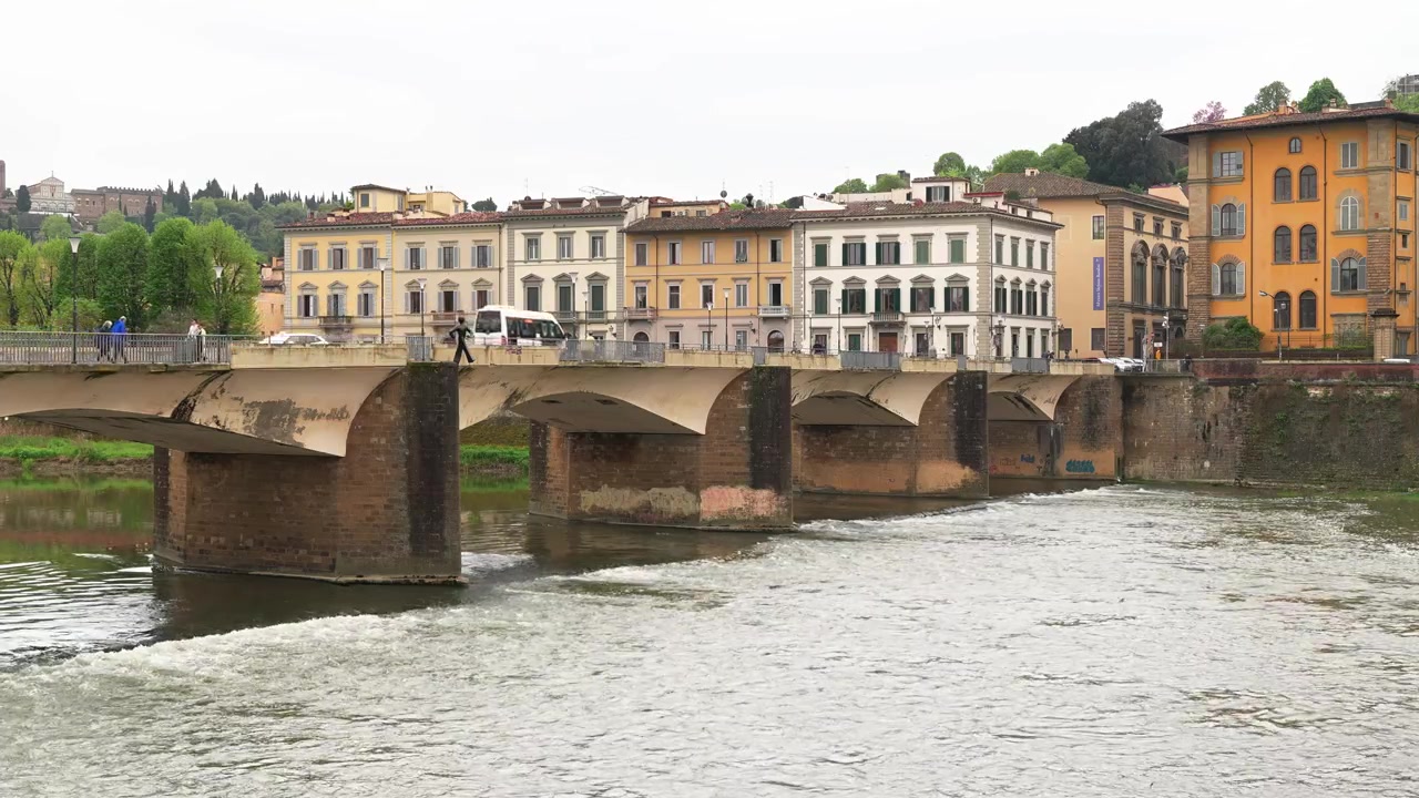 Ampia inquadratura del fiume Arno a Firenze e del suo ponte storico, con eleganti palazzi rinascimentali, pedoni, l'acqua che scorre e una luce naturale soffusa che creano un autentico scorcio della città italiana.