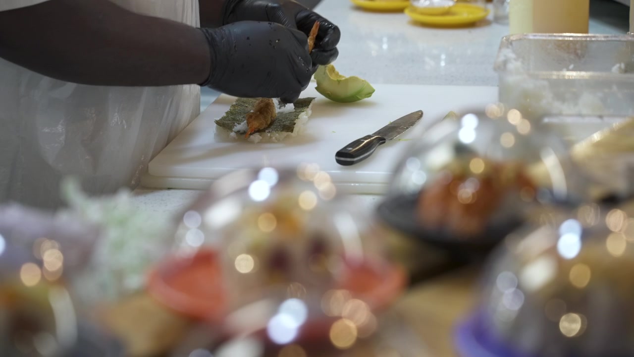 Des mains gantées assemblent un rouleau de sushi à l'avocat sur une planche à découper tandis que des sushis déjà préparés passent au premier plan