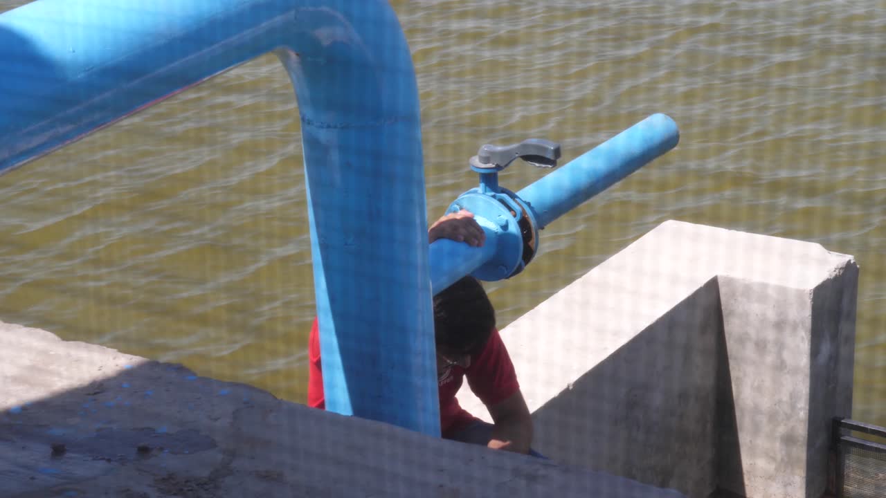 toma en ángulo alto sobre un hombre local sentado junto a un gran grifo azul de un sistema de rociadores de riego de pivote central a lo largo de la orilla del río en punjab durante el día