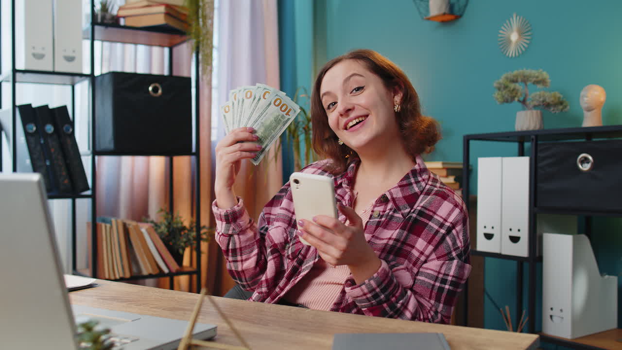 Mujer independiente rica y emocionada mira a la cámara sosteniendo dinero ingresos ganancias dinero en efectivo y teléfono inteligente
