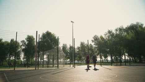 Dos-Jugadores-De-Baloncesto-Masculinos-Caminando-En-Una-Cancha-De-Baloncesto-Al-Aire-Libre,-Mientras-Conversan-Juntos-Y-Uno-De-Ellos-Lanza-La-Pelota