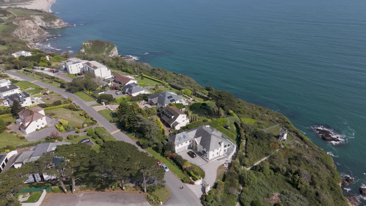 Aerial pan along rugged cliff coastline showcasing modern white luxury homes perched above deep blue waves, manicured lawns, and sweeping ocean vista