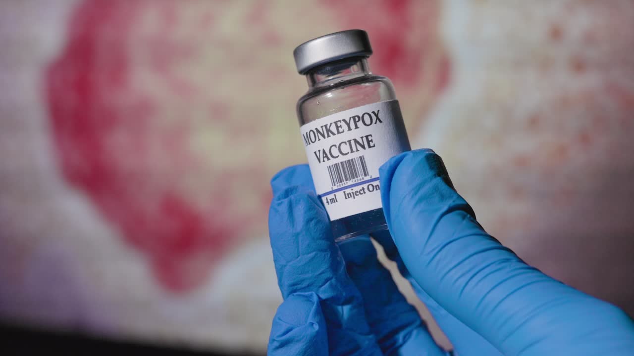 doctor holding a vial of monkeypox vaccine in front of a monitor with the monkeypox virus displayed