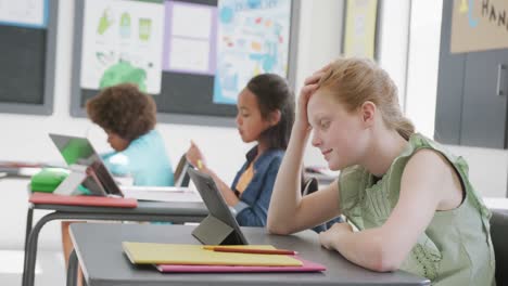 video de una escolar caucásica frustrada sentada en un escritorio usando una tableta en una clase diversa, espacio de copia