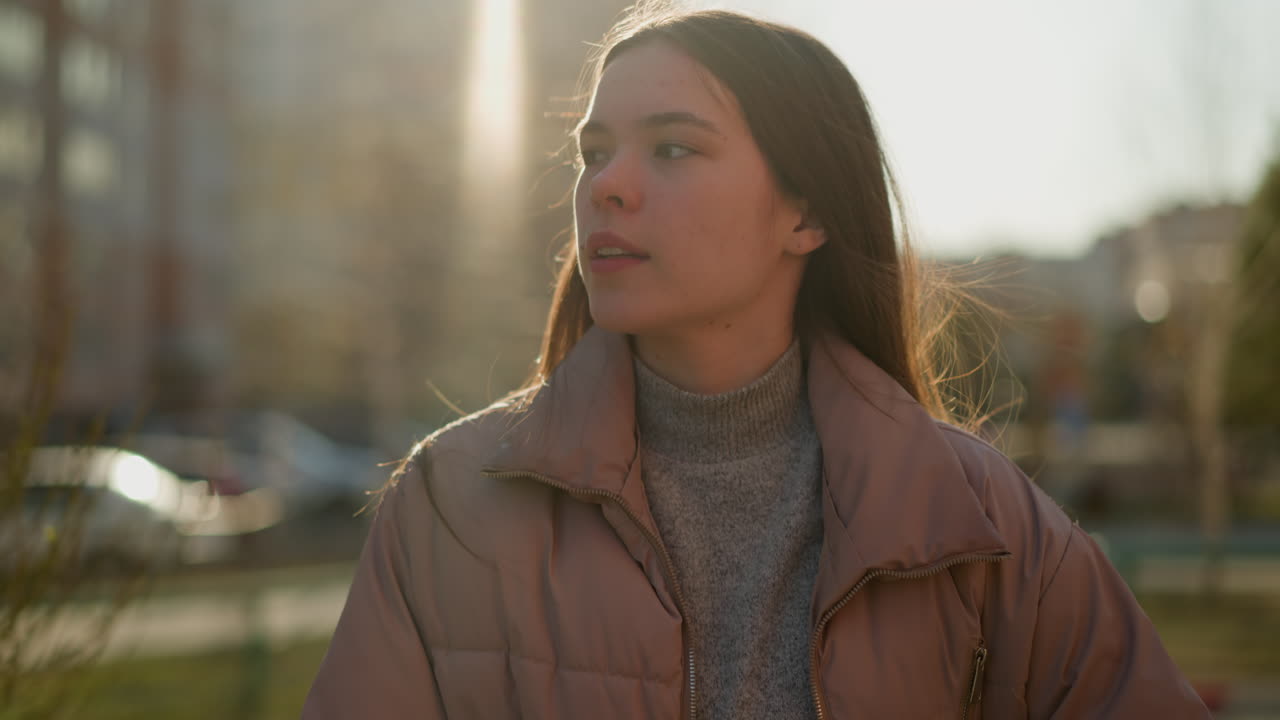 Front view of a girl walking through an urban area at sunset, wearing a peach jacket and a grey inner shirt. The sunlight softly illuminates her face as she gazes ahead with a calm expression