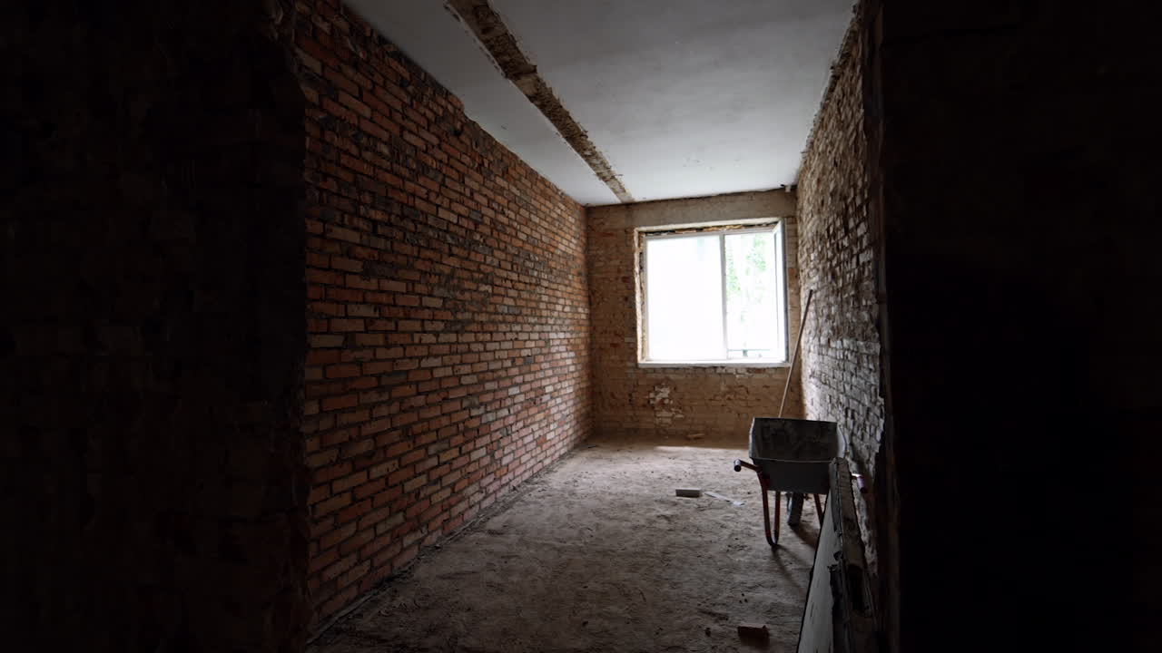 Long room with brick walls and one window. Wheelbarrow and a stick are at the wall. Building renovation.