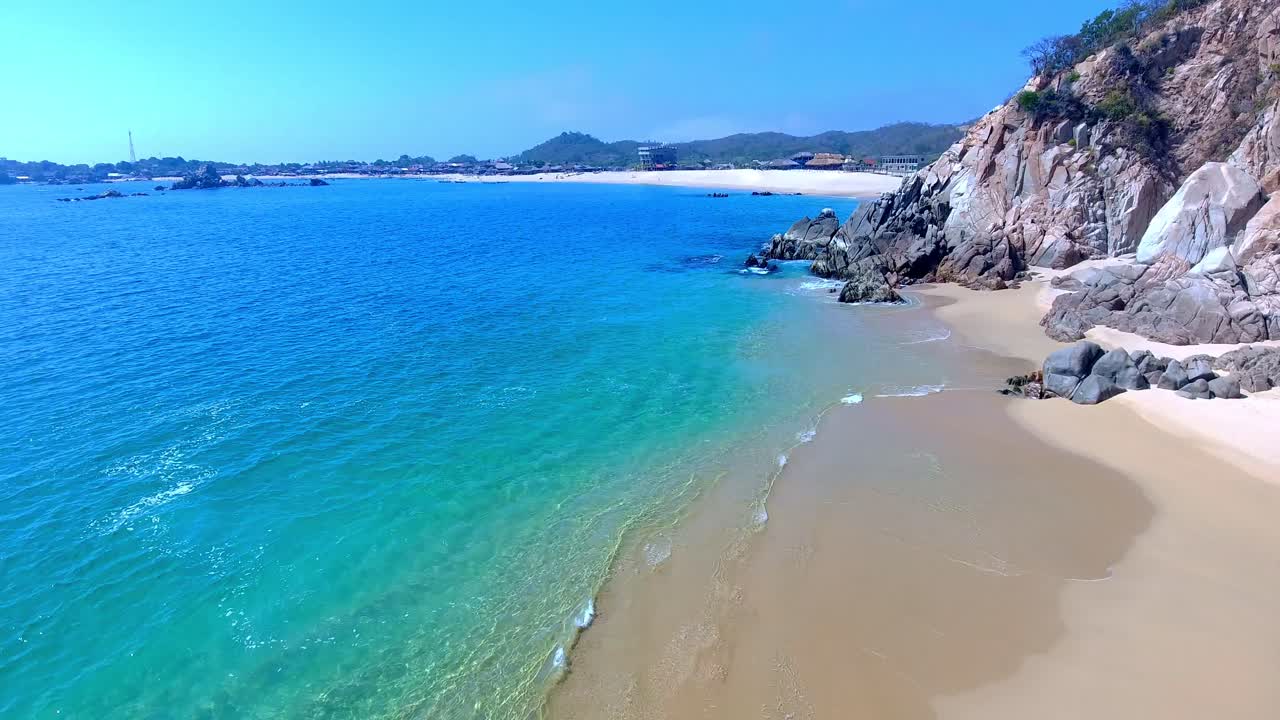 Vista aérea frontal e em ângulo baixo da praia, com pequenas ondas quebrando perto de uma costa rochosa de águas cristalinas.