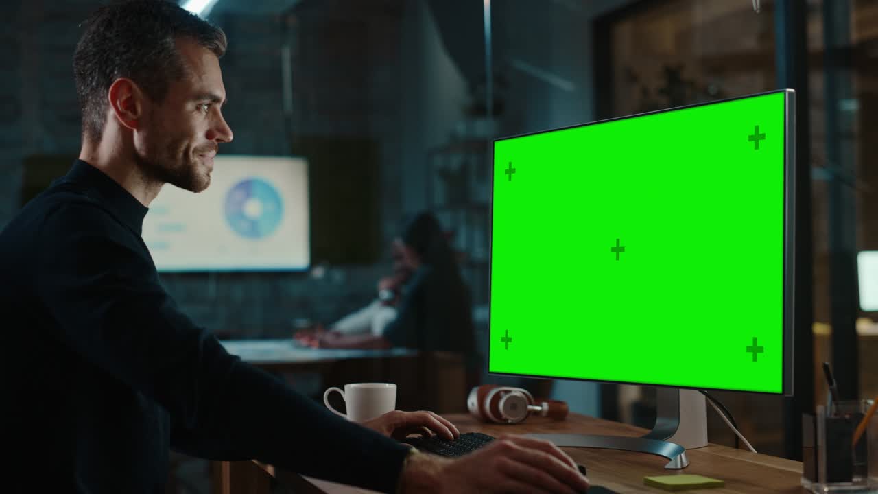 Young Handsome Specialist Working on Desktop Computer with Green Screen Mock Up Display in a Busy Creative Office with Colleagues. Male Manager with Trimmed Beard is Wearing a Casual Black Jumper.