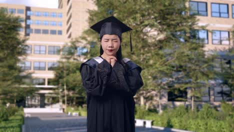 asian woman student graduates in cap and gown pray for something in front of a magnificent university building