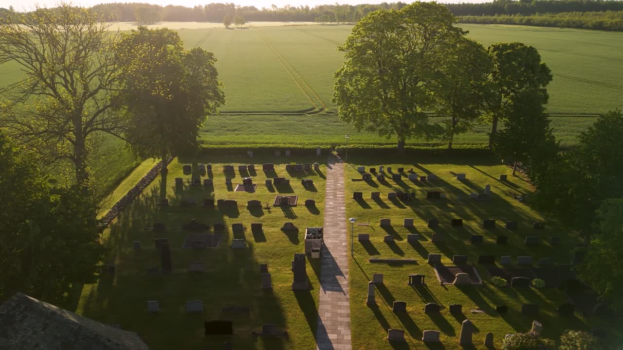 Il drone si muove a destra sopra le scure lapidi di un cimitero che contrastano con un luminoso prato agricolo illuminato dal sole a Mellerud, Svezia, durante il tramonto.