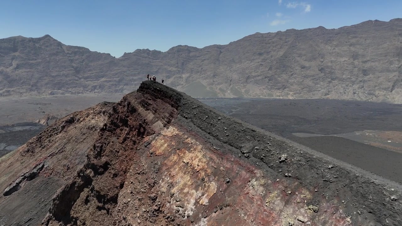 High-altitude drone shot of a group of trekkers standing on the narrow, rugged crater rim of Pico do Fogo