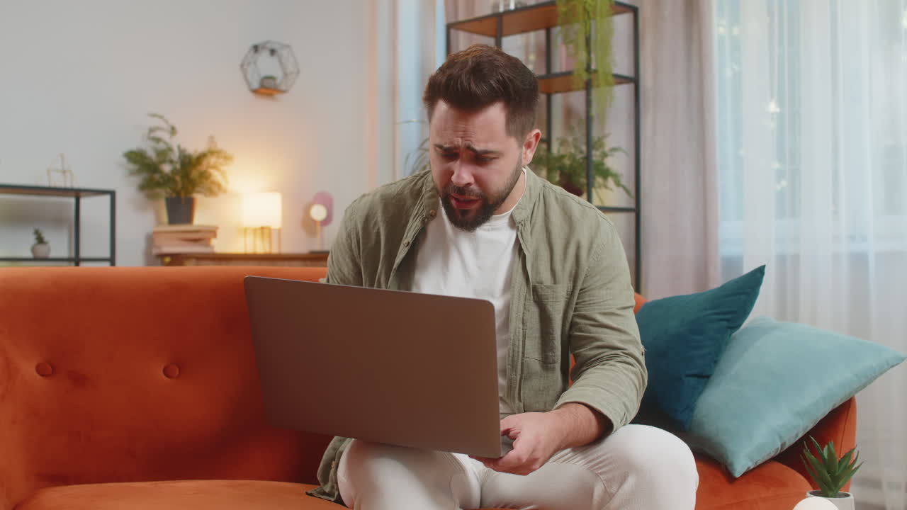 Upset young caucasian man freelancer using laptop engages in heated conversations sitting on sofa