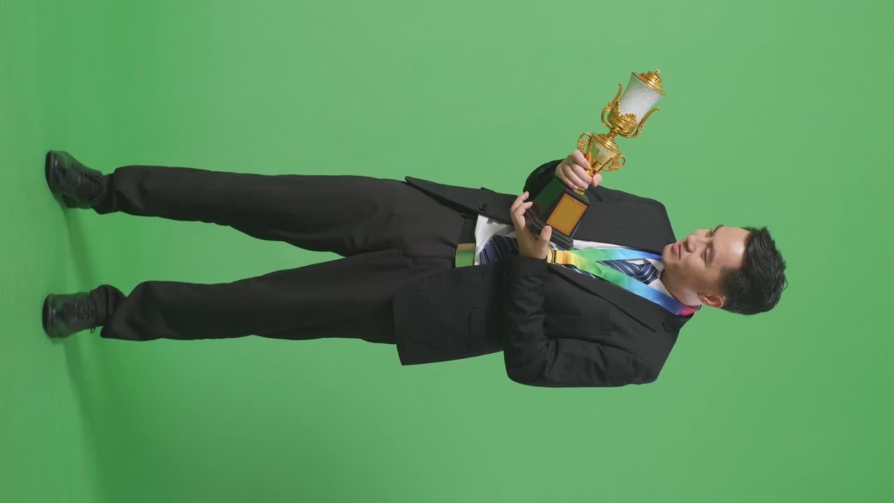 옷과 타이를 입고 황금 메달을 들고 있는 아시아 사업가 한 명이 손에 들고 있는 황금 트로피를 보고 '와우'라고 말하며 스튜디오의 녹색 스크린 배경에서 첫 번째 우승자로서 행복하다.